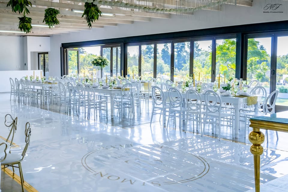 Dakalo Glass Venue set for a wedding, long white tables and ghost chairs against floor-to-ceiling garden windows